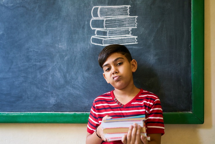 Concept on blackboard at school. Young people, student and pupil in classroom. Sad and bored hispanic boy in class. Portrait of male child looking at camera, holding books